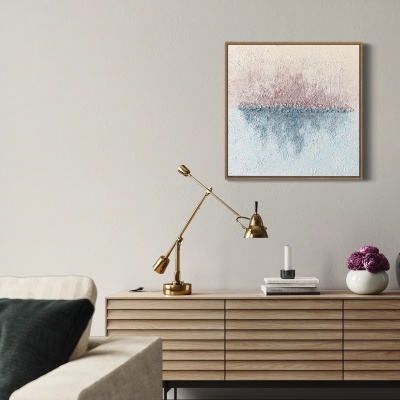 Sala com cómoda de madeira clara, lâmpada dourada, vela, vaso com flores e pintura abstrata na parede.