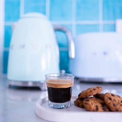Copo de café expresso e bolachas sobre bandeja branca com chaleira e torradeira ao fundo