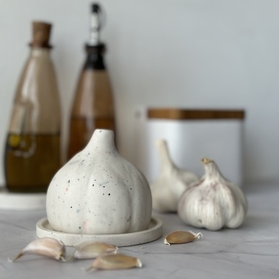 Cabeça de alho branca numa base branca com dentes de alho sobre bancada cinza, garrafas e frasco ao fundo
