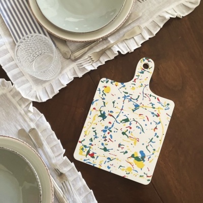 Tábua de cortar colorida ao lado de loiça branca e copos numa mesa de madeira