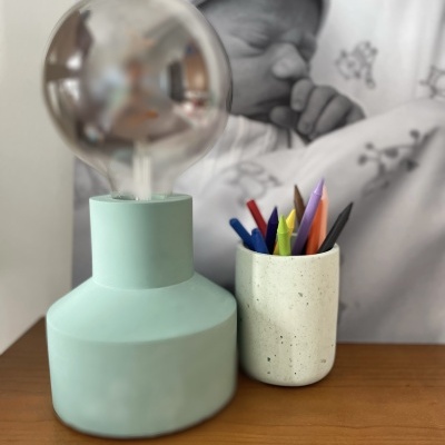 Lâmpada verde pastel e copo de cimento com canetas coloridas numa mesa de madeira