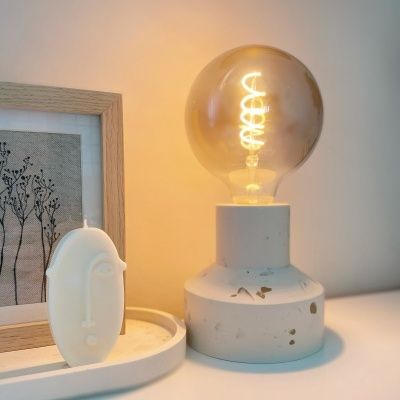 Abajur de base branca com lâmpada de filamento, vela decorativa branca e quadro com ilustração de plantas num ambiente interior.