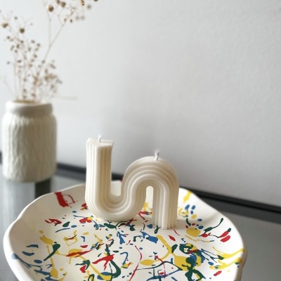 Vela branca em forma de L e U num prato colorido sobre uma mesa com vaso branco e ramo seco
