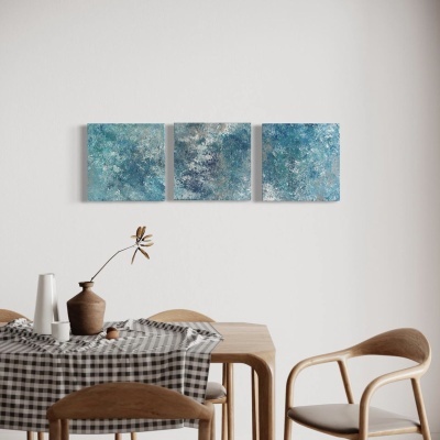 Três quadros azuis abstratos numa parede branca acima de uma mesa de madeira com toalha xadrez e cadeiras de madeira