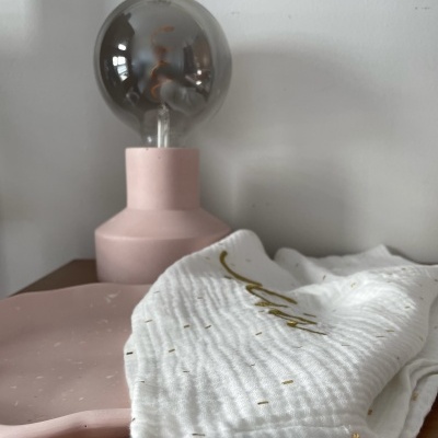 Lâmpada de mesa rosa com lâmpada de vidro fumado e tecido branco com detalhes dourados sobre uma superfície de madeira