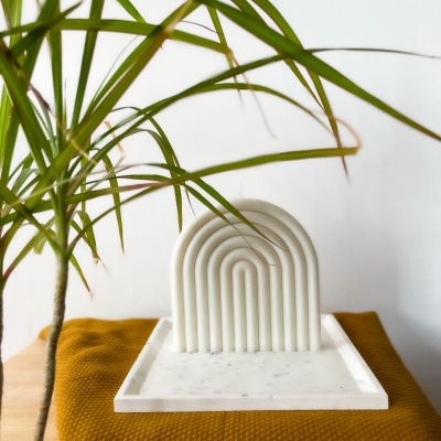 Suporte decorativo branco com padrão de arcos em cima de tecido mostarda e planta ao lado