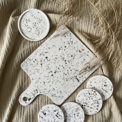 Tábua de cortar e bases redondas em material marmoreado branco com manchas escuras sobre tecido bege com ramos secos