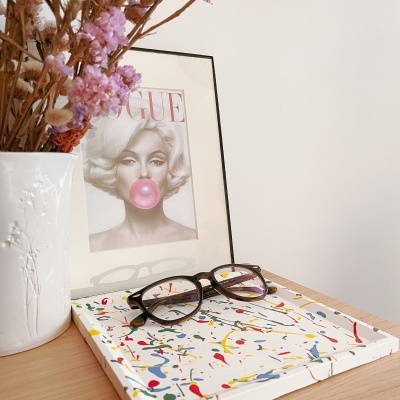 Moldura com capa da VOGUE, vaso branco com flores secas, livro colorido e óculos sobre mesa de madeira clara