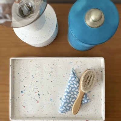 Objetos decorativos numa mesa de madeira incluindo uma lâmpada, recipiente azul, tabuleiro com padrão pontilhado, pano azul estampado e escova de madeira.