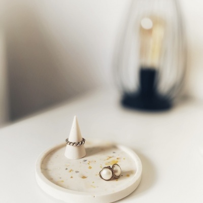 Anel prateado torcido e brincos de pérola sobre base de mármore branca com manchas douradas e candeeiro ao fundo