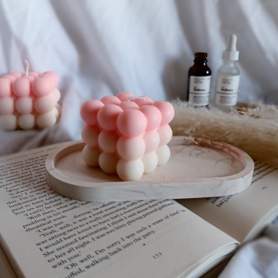 Vela cubo rosa e creme em prato de mármore com frascos e livro aberto ao fundo