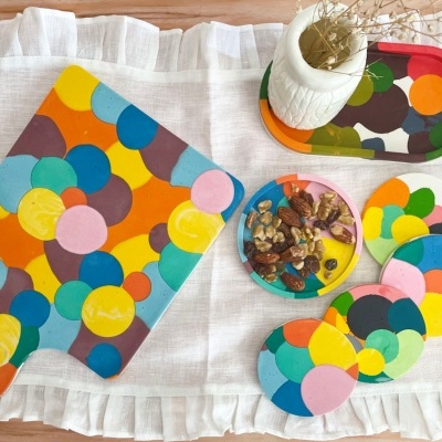 Tábua de cortar, tabuleiro, prato de nozes e descansos de copo com padrão de círculos multicoloridos sobre pano branco com babados