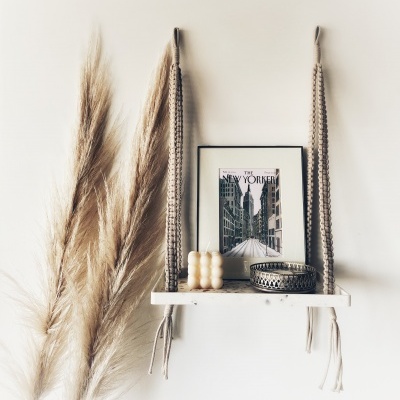 Prateleira suspensa com vela, caixa decorativa e quadro de cidade na parede branca com plantas secas ao lado.