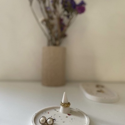 anel dourado, brincos pérola em prato branco manchado com vaso bege e flores secas desfocado ao fundo