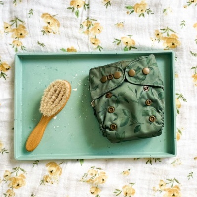 Fralda reutilizável verde com folhas escuras e escova de cerdas com cabo de madeira numa bandeja verde clara