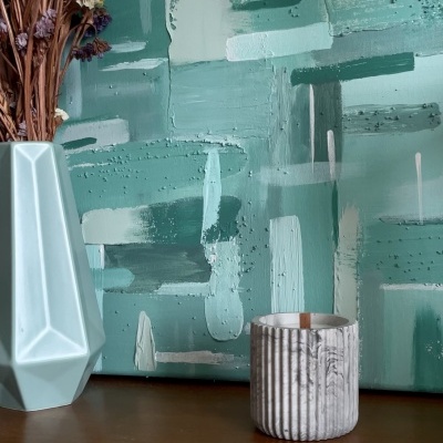 Vaso azul com flores secas e vela em suporte cerâmico sobre mesa de madeira com pintura abstrata verde ao fundo