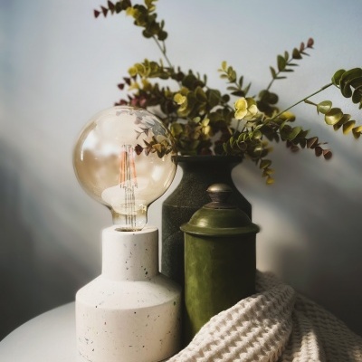 Vaso verde com flores, candeeiro branco com lâmpada e frasco verde sobre mesa com manta creme