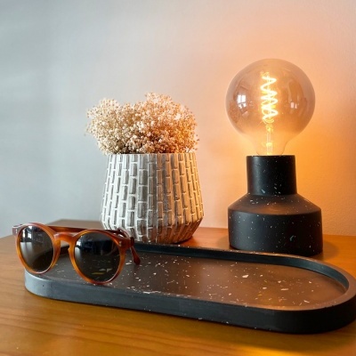 Mesa de madeira com suporte cinzento, óculos de sol, vaso com flores secas e lâmpada iluminada.