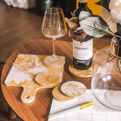 Mesa redonda com vinho ASSOBIO, copo, bases e tábua marmoreada, jarro com flores e papel com caneta.