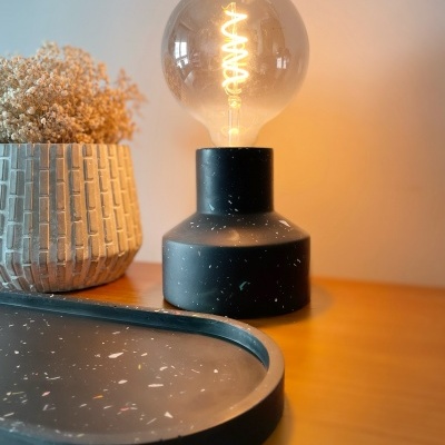 Abajur preto com lâmpada vintage, vaso cinza geométrico com flores secas e bandeja preta sobre mesa de madeira clara