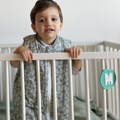Criança num berço branco com saco de dormir verde e etiqueta 'M' pendurada
