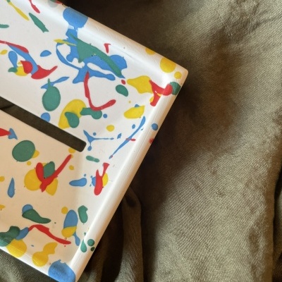 Objeto retangular branco com padrão colorido sobre tecido verde-oliva