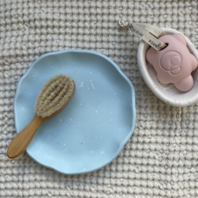 Escova de madeira com cerdas naturais, prato azul claro e sabonete rosa sobre tecido branco texturizado