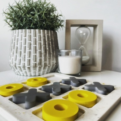 Jogo da velha amarelo e cinzento em tabuleiro branco com planta, vela e ampulheta ao fundo