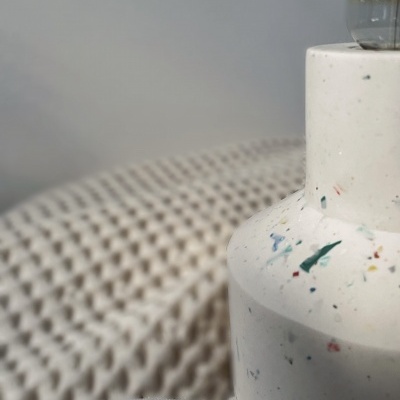 Lâmpada de mesa branca com manchas coloridas e luz visível, sobre superfície texturizada cinza-claro