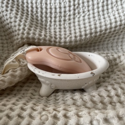 Saboneteiro branco em forma de banheira com sabonete rosa claro sobre tecido branco texturizado