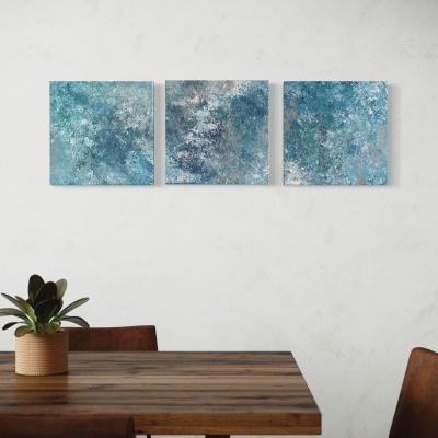 Três quadros abstratos azuis pendurados numa parede branca sobre mesa de madeira com cadeiras e planta.