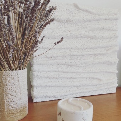 Vaso branco com flores secas, painel texturizado branco e vela branca numa mesa de madeira.