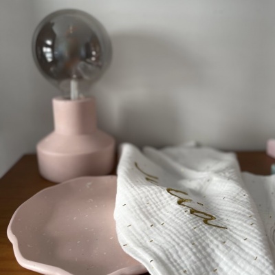 Lâmpada decorativa rosa, toalha branca com escrita dourada 'Fuller' e prato rosa sobre superfície de madeira