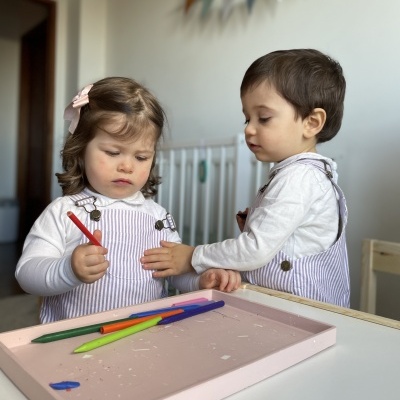 Duas crianças em ambiente interior com bandeja rosa e lápis coloridos sobre mesa branca