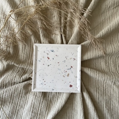 Prato quadrado de cerâmica com padrão terrazzo sobre tecido bege canelado e elementos decorativos secos