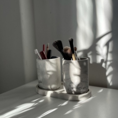 Suportes cilíndricos de mármore branco com pincéis de maquilhagem, sobre base oval e superfície branca com sombras naturais