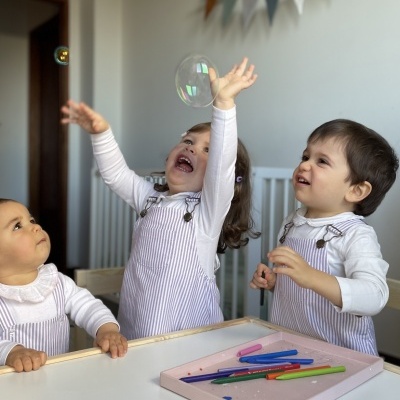 Três crianças pequenas brincando à volta de mesa com bandeja de lápis de cores, bolha de sabão no ar, fundo com berço e decoração de parede
