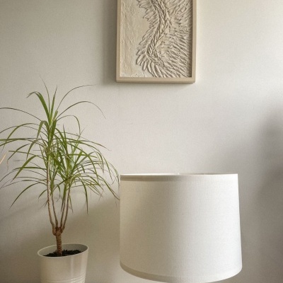 Planta em vaso branco, candeeiro de madeira com cúpula branca e quadro decorativo em relevo numa parede branca