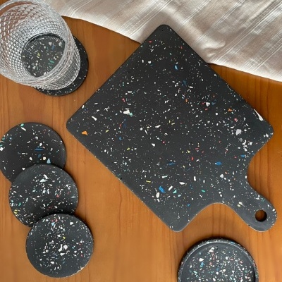 Tábua de corte e bases para copos pretas com padrão marmoreado colorido sobre mesa de madeira e pano branco.