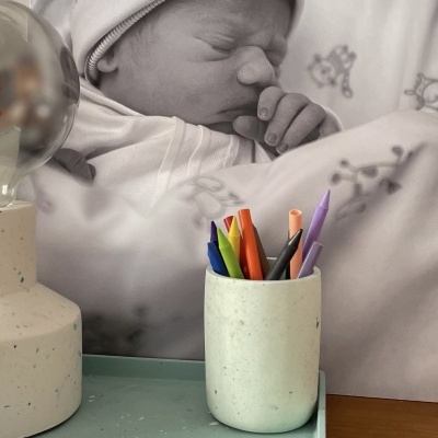 Suporte de canetas com lápis coloridos numa bandeja verde e lâmpada com base creme em frente a foto de bebé a dormir em preto e branco