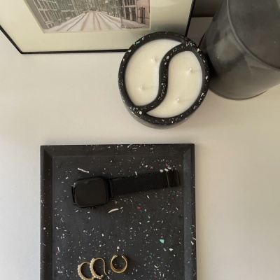 vela dupla branca em recipiente preto, relógio inteligente preto e anéis dourados em tabuleiro preto manchado, jarra cinza e quadro urbano