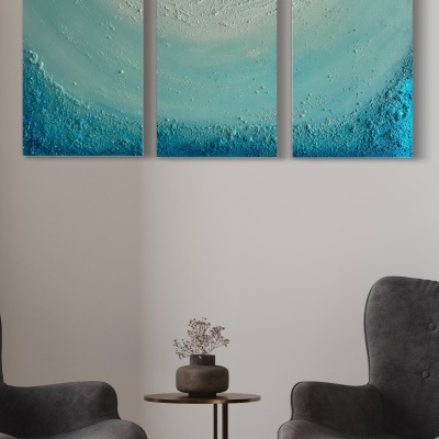 Conjunto de três quadros abstratos azuis e verdes numa parede branca entre duas poltronas cinzentas e uma mesa com vaso.