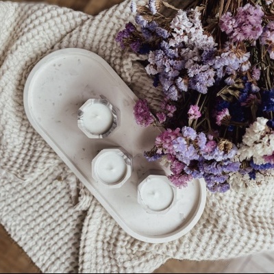Três velas brancas em suportes hexagonais de cimento numa bandeja oval sobre um cobertor creme com flores secas coloridas ao lado