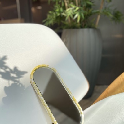 Espelho oval com moldura amarela e branca entre duas cadeiras brancas e planta verde em vaso cinza