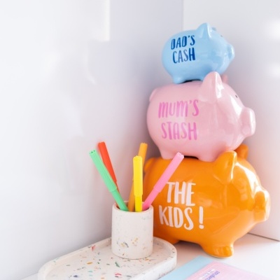 Três mealheiros coloridos empilhados com texto, canetas coloridas em recipiente cinzento e base branca com manchas coloridas