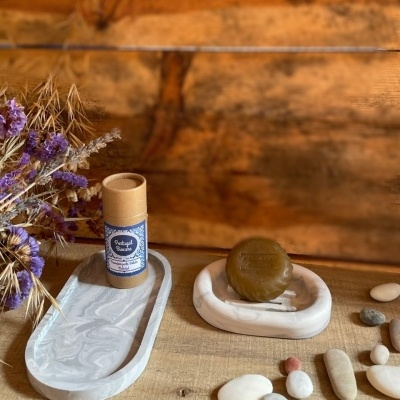 Sabonete redondo castanho em saboneteira branca com embalagem cilíndrica e flores secas ao lado.