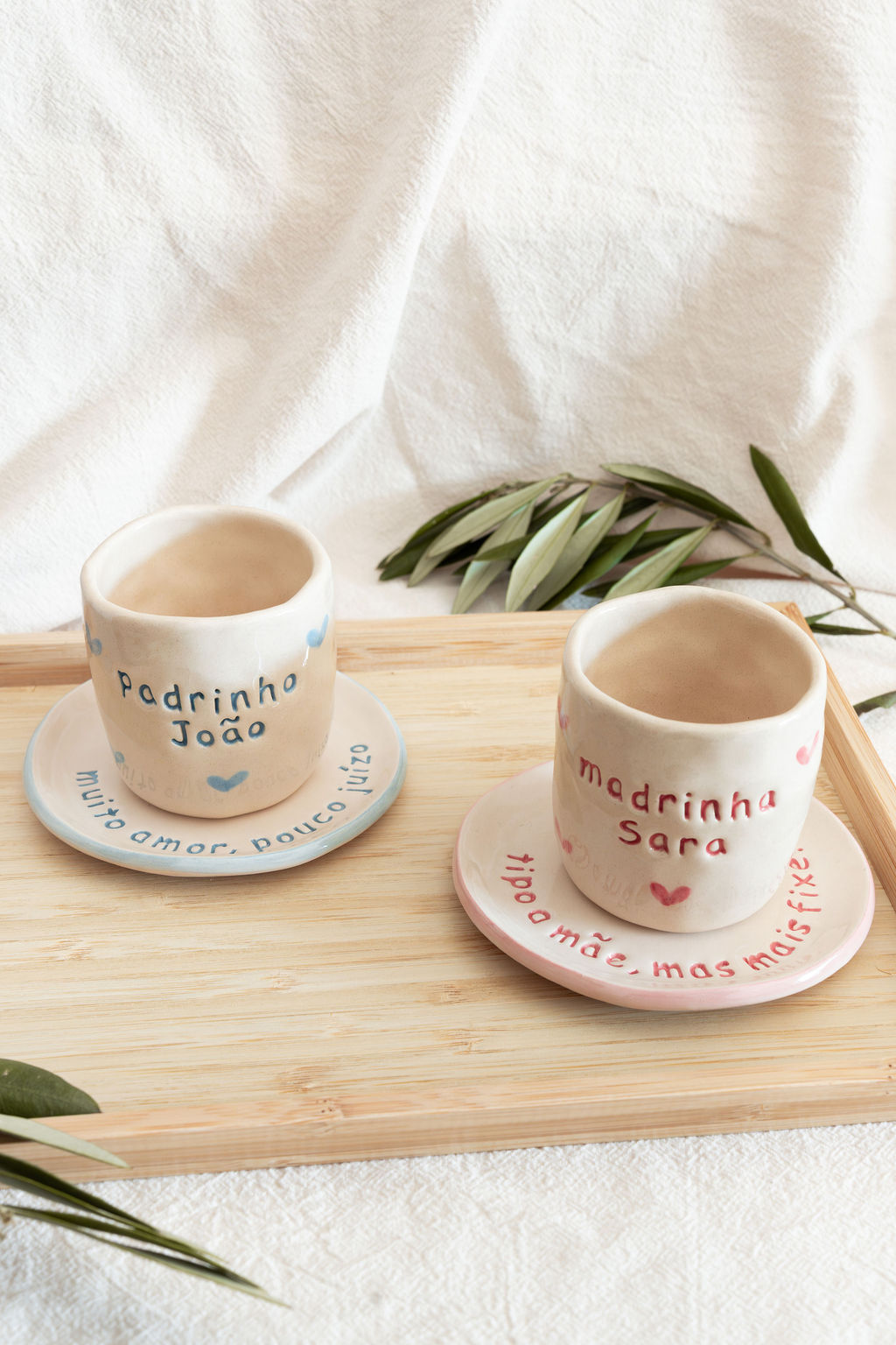 Duas chávenas de cerâmica com pires, uma azul com texto Padrinho João e outra rosa com texto Madrinha Sara sobre bandeja de madeira.
