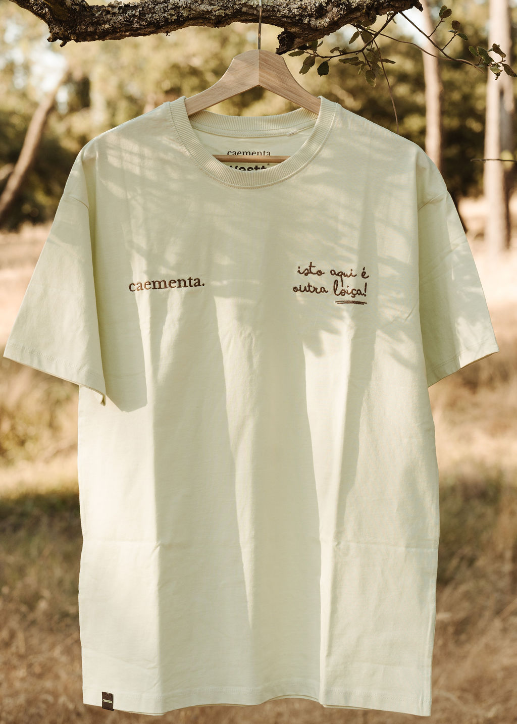 T-shirt creme com texto bordado castanho pendurada em ramo de árvore