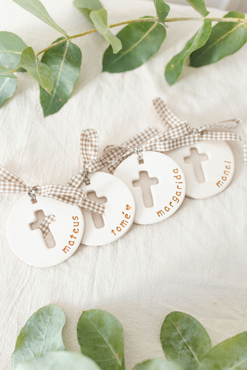 Ornamentos redondos de cerâmica branca com cruz e nomes, com laços xadrez castanhos e brancos, sobre tecido creme e folhas verdes