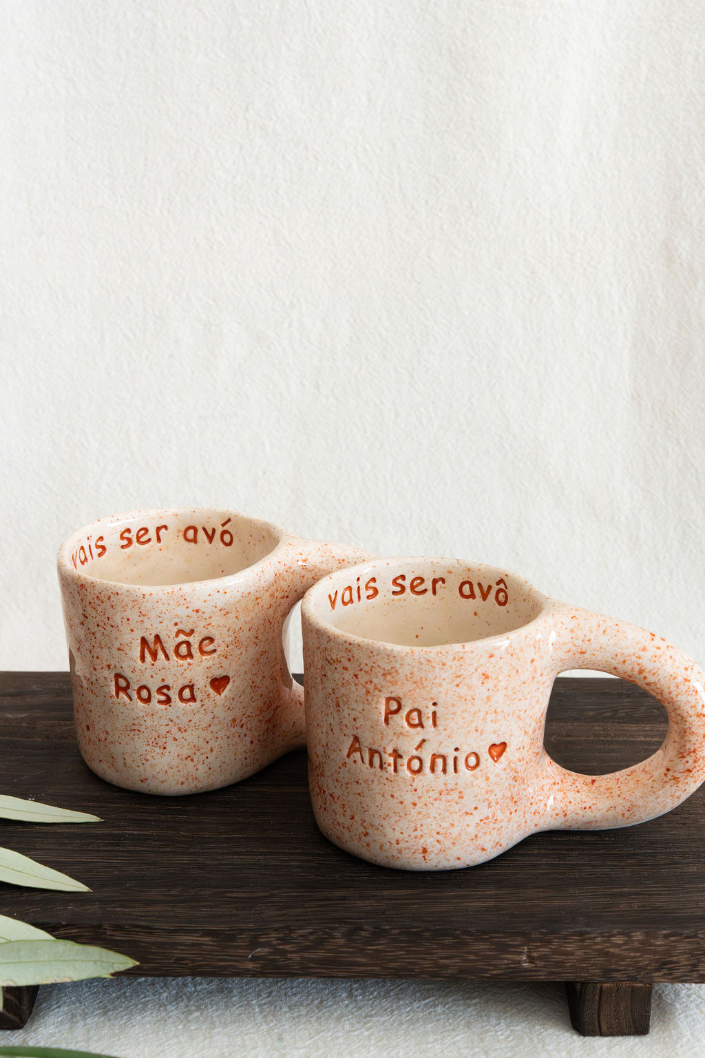 Duas canecas cerâmicas creme com salpicos laranja e texto em relevo sentado numa tábua de madeira.